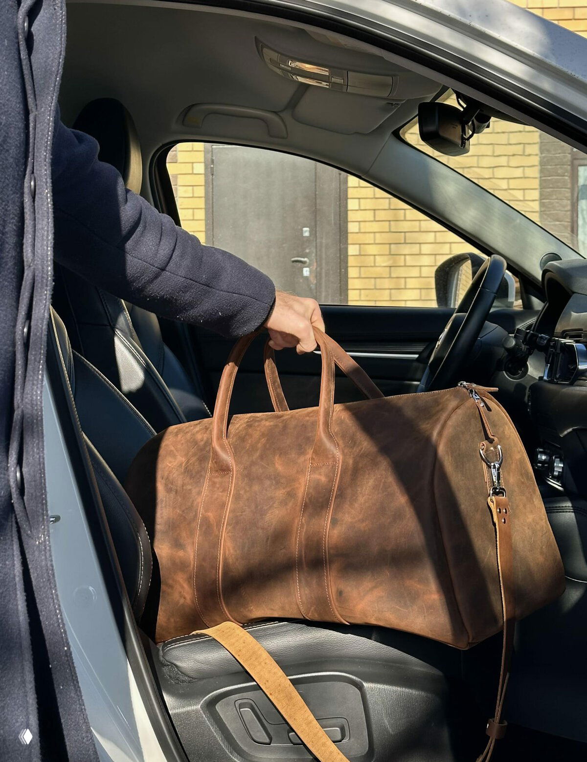 Коричневый дорожный саквояж из фактурной кожи в салоне автомобиля, сверху видна рука человека, подчеркивающая размеры и практичность изделия.