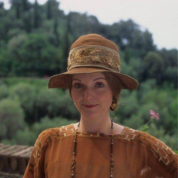 Женщина в шляпе улыбается на фоне зелёного итальянского пейзажа — спокойный кадр из весенней экранизации, тёплые тона.