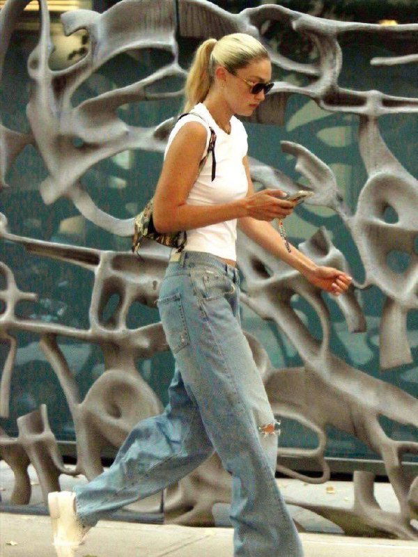 Повседневный снимок: женщина в белой футболке и мешковатых джинсах идёт по улице, опустив взгляд на телефон.