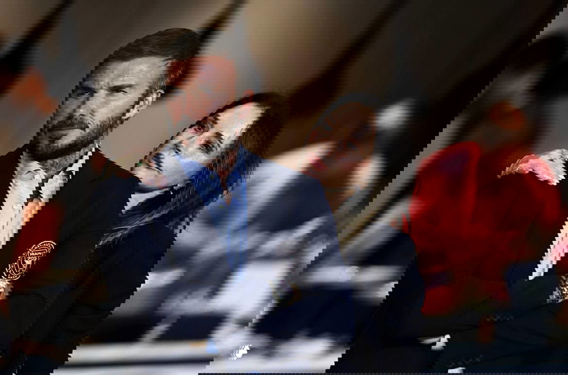 Дэвид и Виктория Бекхэм вместе на общественном мероприятии; Виктория опирается на плечо мужа, на фоне размытие гостей и интерьер зала.