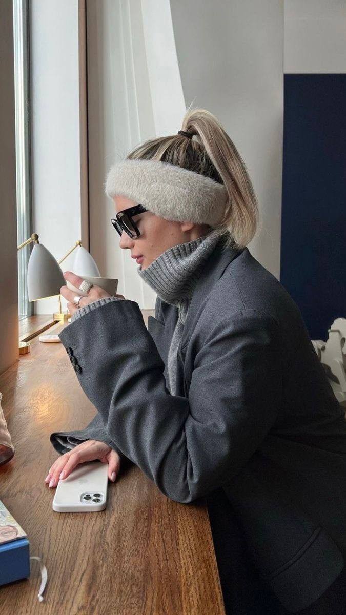 Профиль женщины в светлой пушистой повязке-наушнике и тёплом пальто, сидит у окна с чашкой на столе, образ в городском кафе.