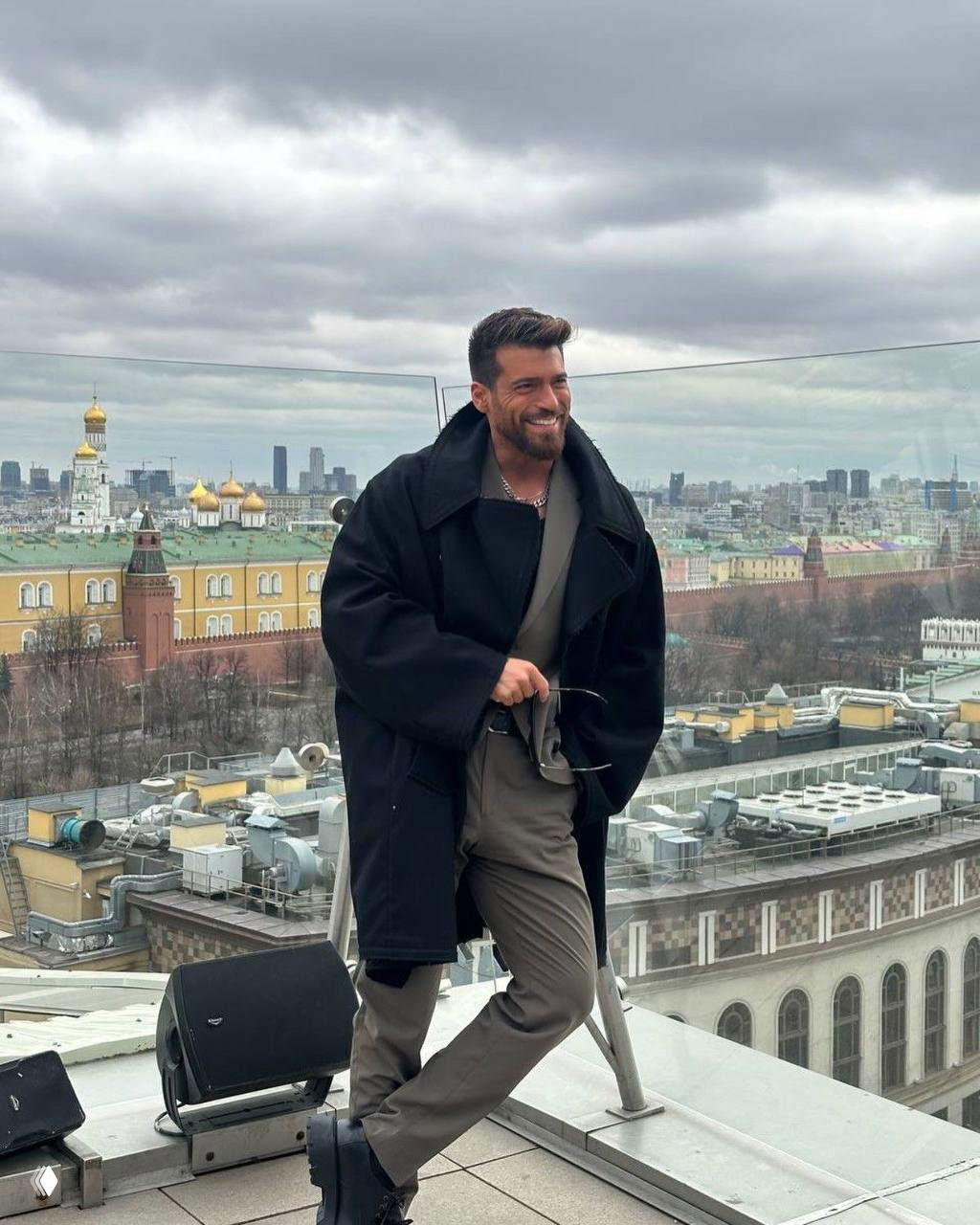 Джин Яман на крыше в Москве: актёр в пальто улыбается на фоне городского пейзажа и виднеющегося храма с золотыми куполами.