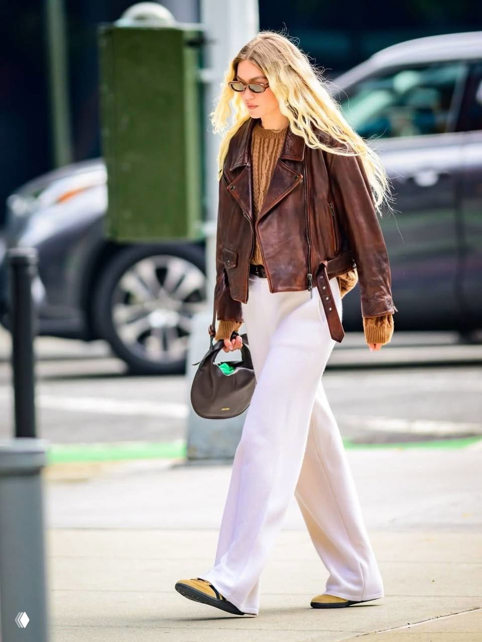 Городской кадр: модель в коричневой кожаной куртке, трикотажном свитере и белых широких брюках идёт по перекрёстку.