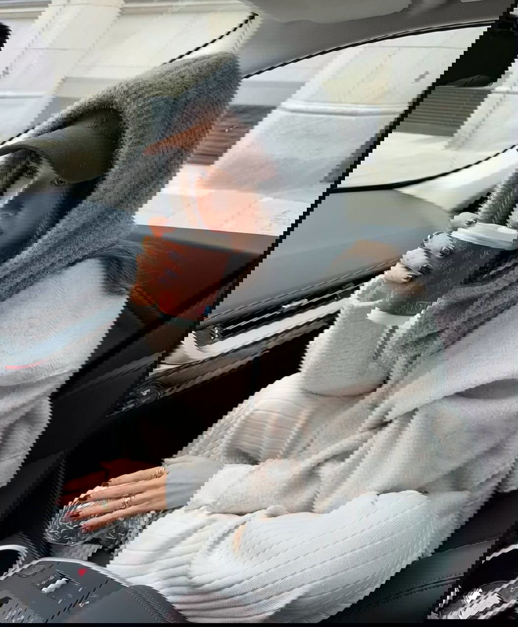 Девушка в автомобиле в объёмном шарфе-капоре и бейсболке, держит стакан кофе — сочетание капора и кепки в повседневном зимнем образе.