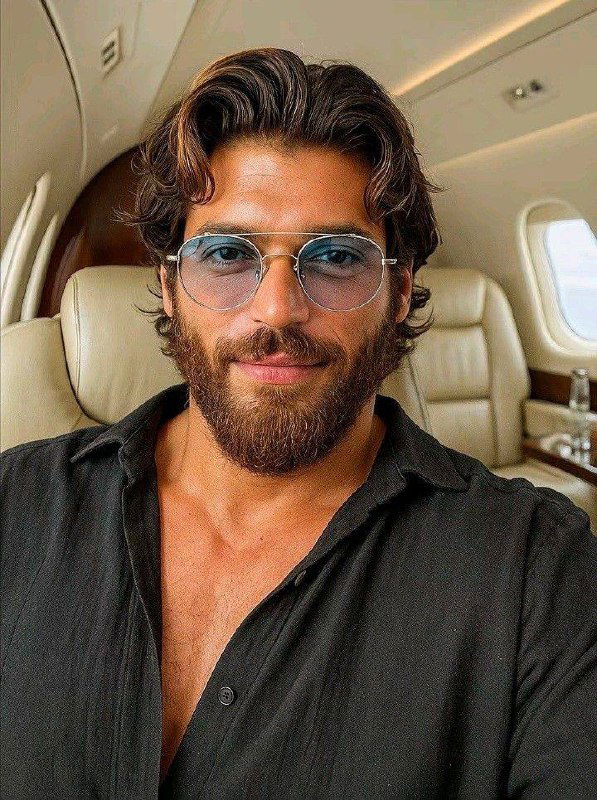 Крупный портрет Джина Ямана в самолёте: он в тёмной рубашке и очках, тёплый свет подчёркивает бороду и улыбку актёра в неформальной обстановке.