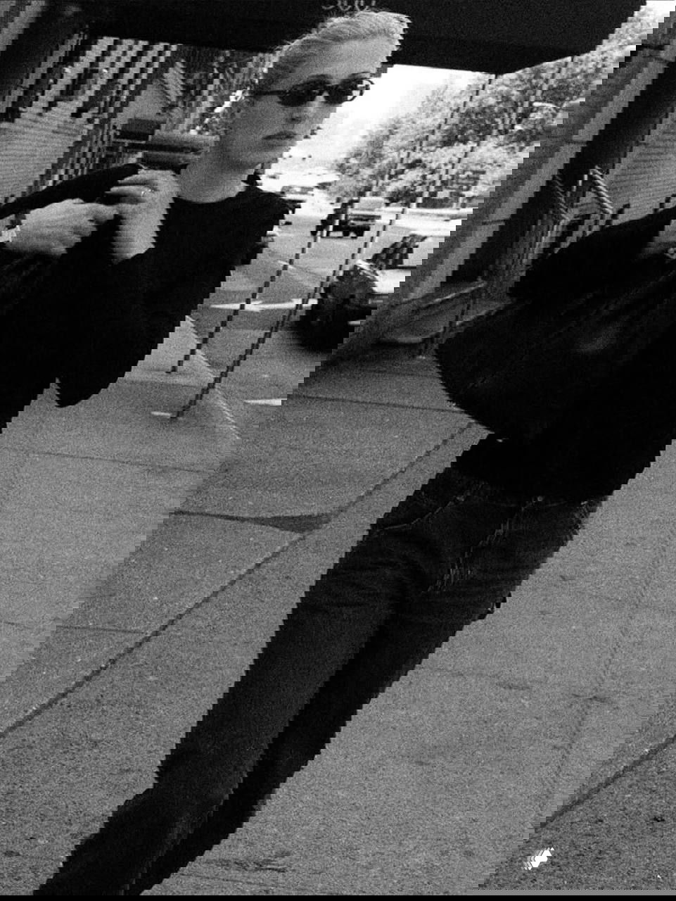 Черно-белая фотография: женщина в темном свитере и джинсах идет по улице, держит большую сумку; фото в документальном, папарацци-стиле.