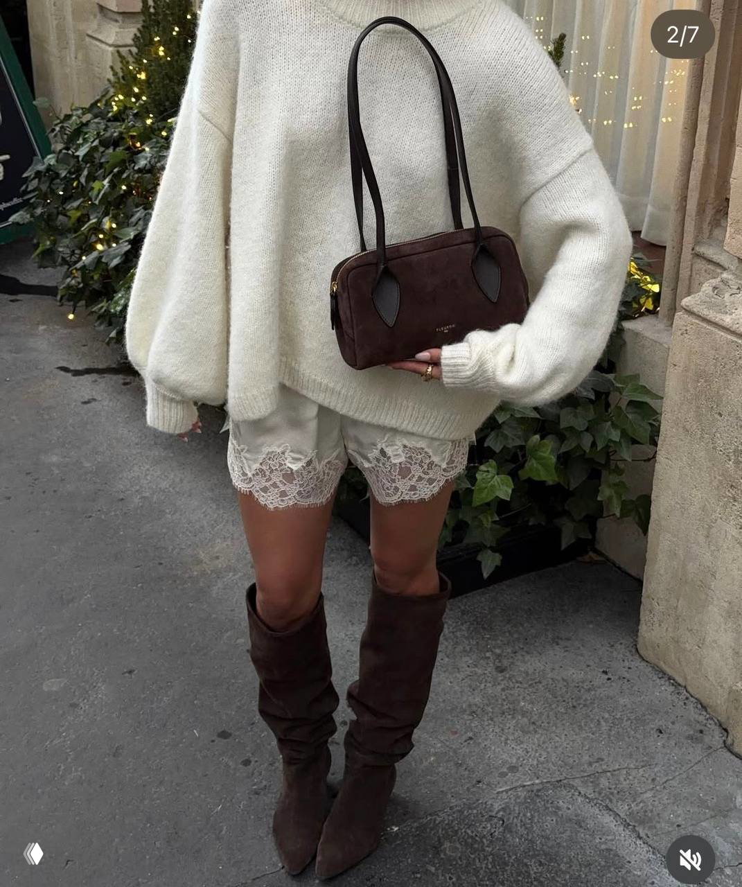 Образ в уютном свитере и кремовых шортах с кружевной отделкой на подоле, в руках — сумка, городской фон и праздничная гирлянда на заднем плане.