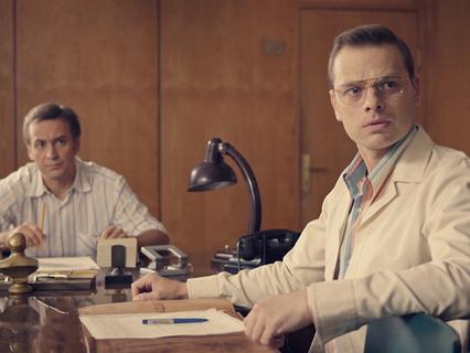 Кадр из ретродетективного сериала: два мужчины сидят за столом в комнате с лампой и бумагами, создавая атмосферу советского кабинета и расследования.