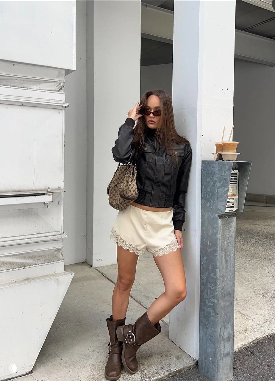 Модель опирается о колонну в кожаной куртке и кремовых шортах с кружевом, образ дополнен высокими сапогами и ретро-сумкой, городская архитектура рядом.