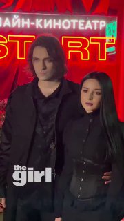 Два гостя в тёмной одежде позируют на фоне неоновой стены с надписями; кадр с красной подсветкой и логотипом СМИ на премьере