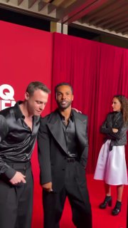 Крупный план двух мужчин в одинаковых черных шелковых рубашках на фоне красной стены с логотипом GQ; сценический кадр с гостями.