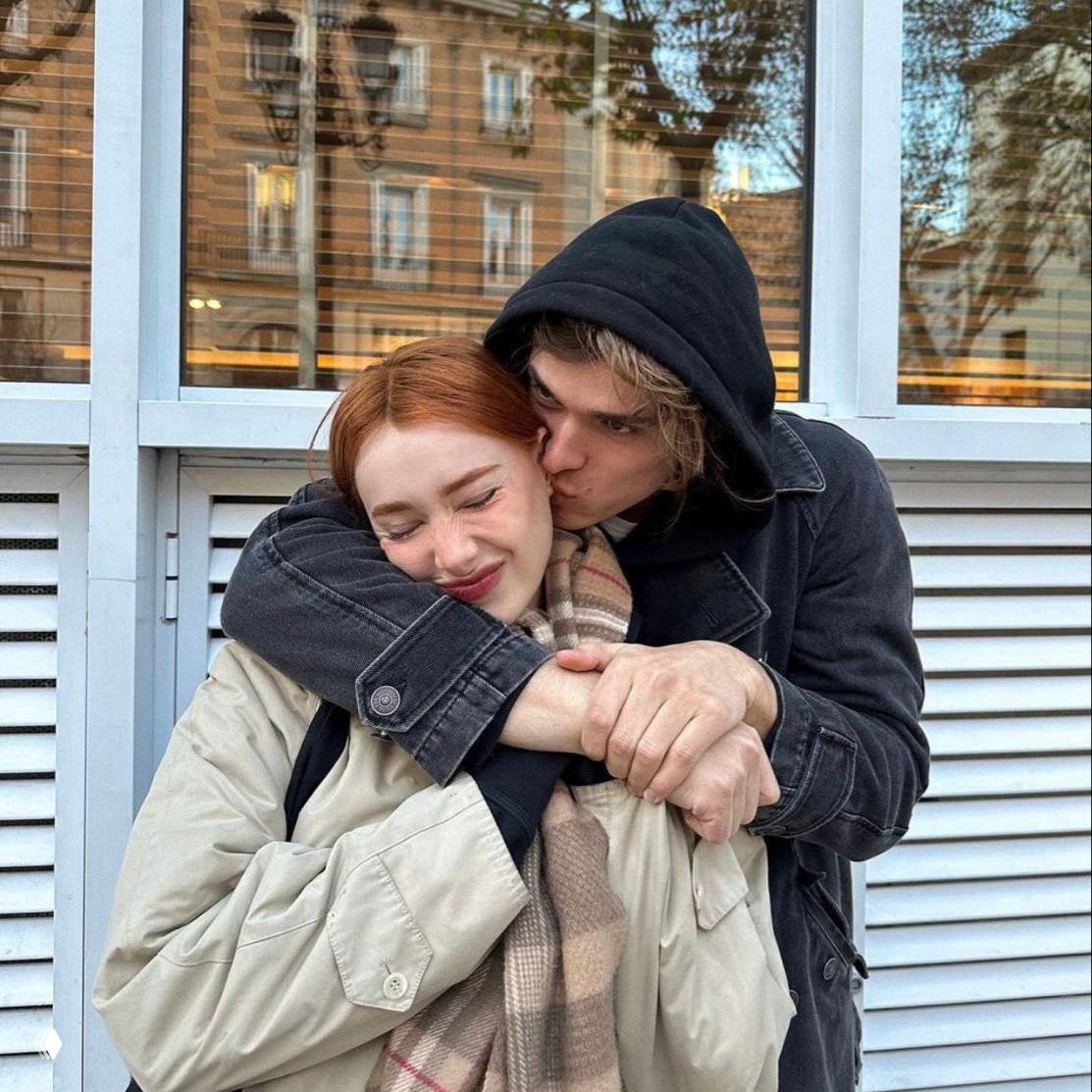 Уличный кадр: пара обнимается у витрины, рыжая девушка улыбается, молодой человек целует её в щёку — романтичный городской образ.