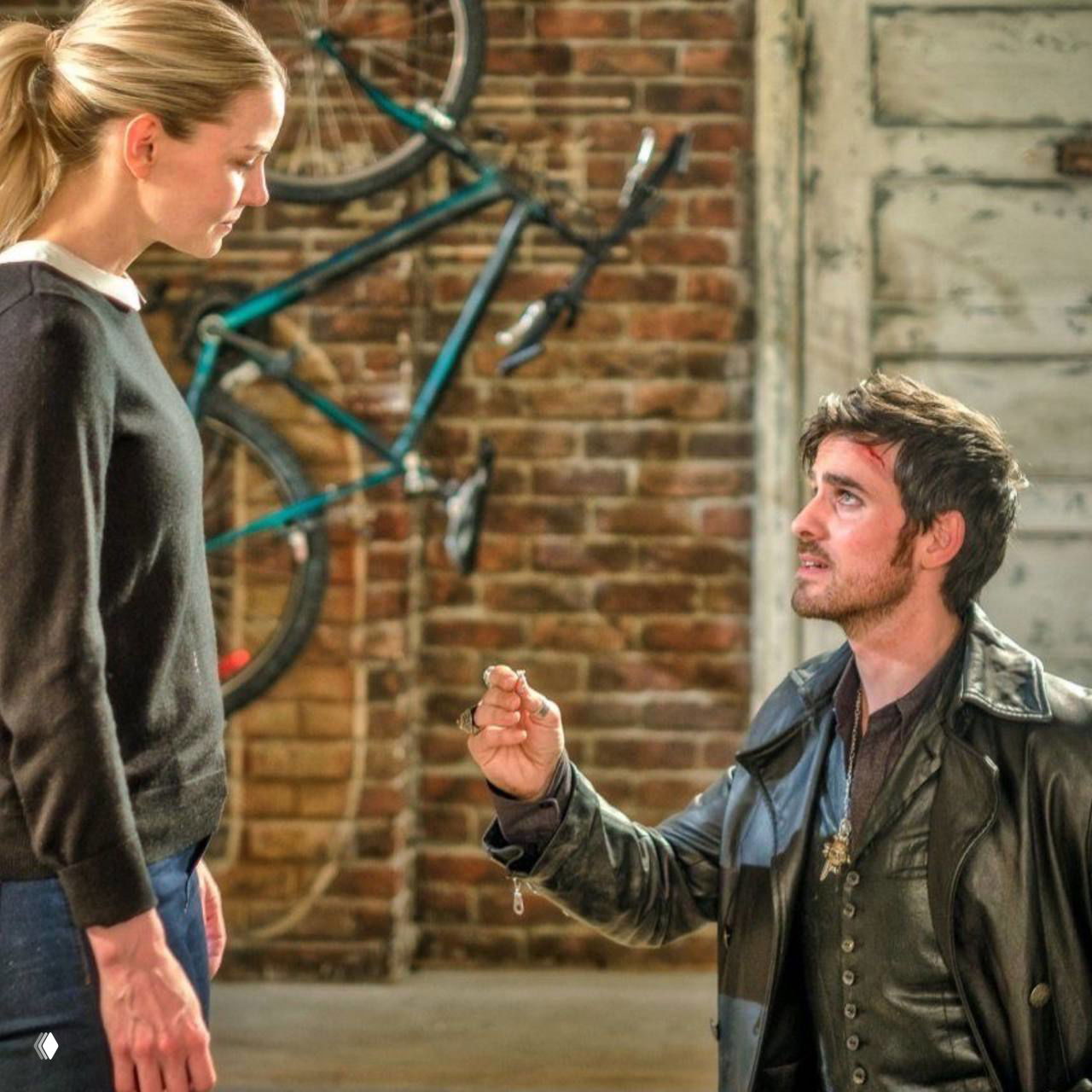 Сцена на складе: Киллиан преклоняется перед Эммой, протягивая ей предмет, в фоне велосипед и кирпичная стена — момент признания и доверия.