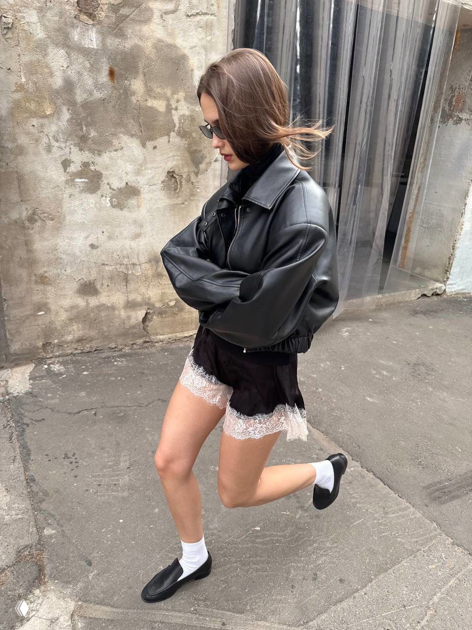 Динамичный кадр: модель в тёмной куртке и чёрных шортах с кружевной кромкой в движении по асфальту, контраст текстур и спортивная обувь.
