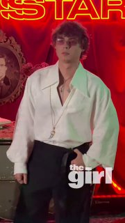 Мужчина в белой рубашке на фоне неоновой вывески и красной декорации с логотипом СМИ; кадр с премьеры, вечерняя сцена.