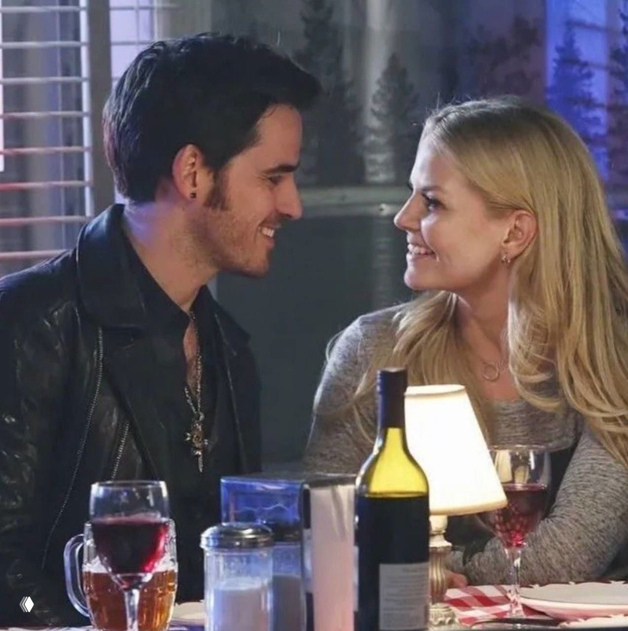 Романтический ужин: Эмма и Киллиан улыбаются друг другу за столом с бокалами вина и бутылкой на переднем плане, тёплая сцена встречи.