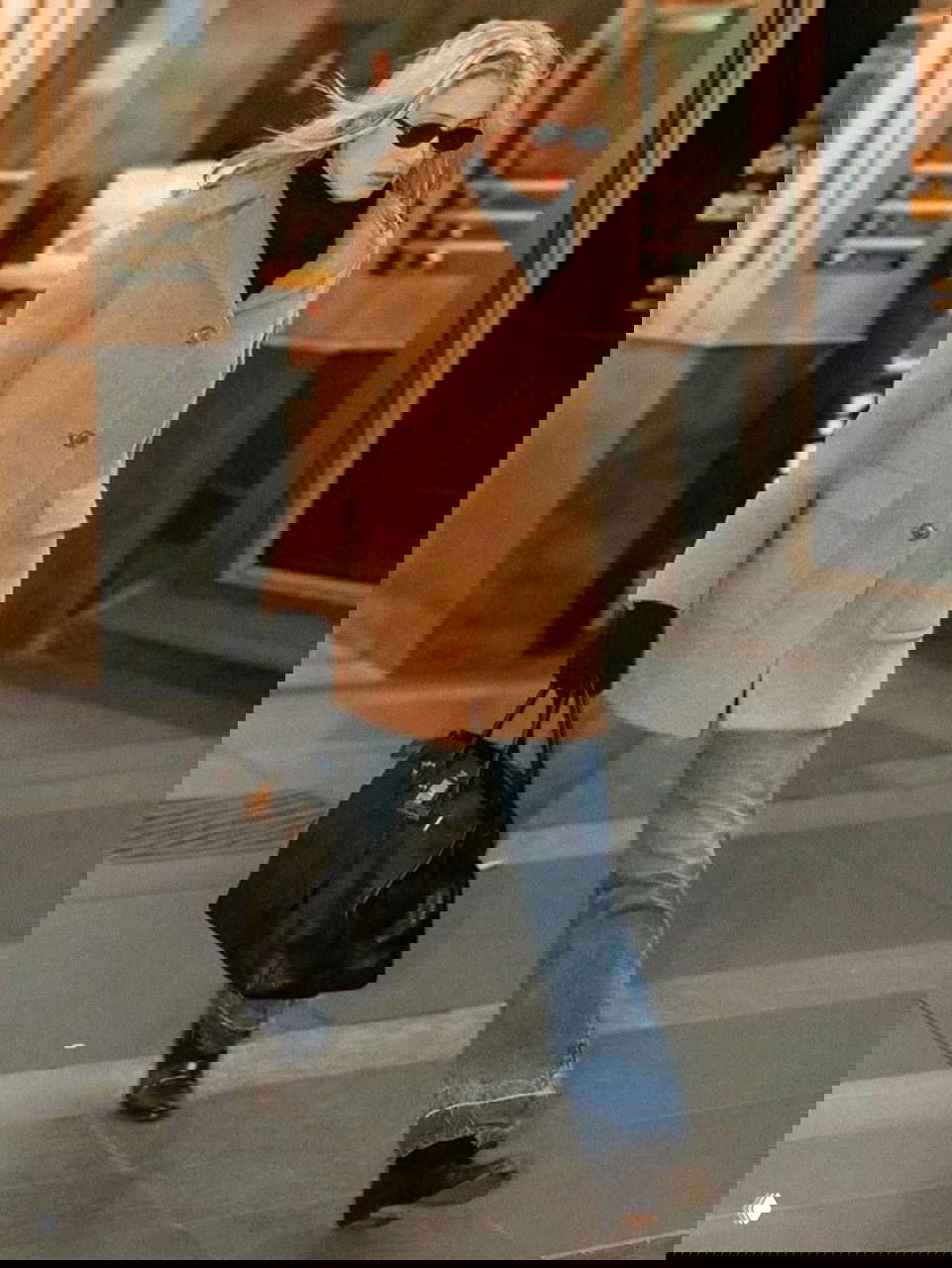 Женщина в бежевом пальто, солнечные очки и джинсы спешит по городским улицам, держа большую кожаную сумку; мягкий теплый цвет кадра.