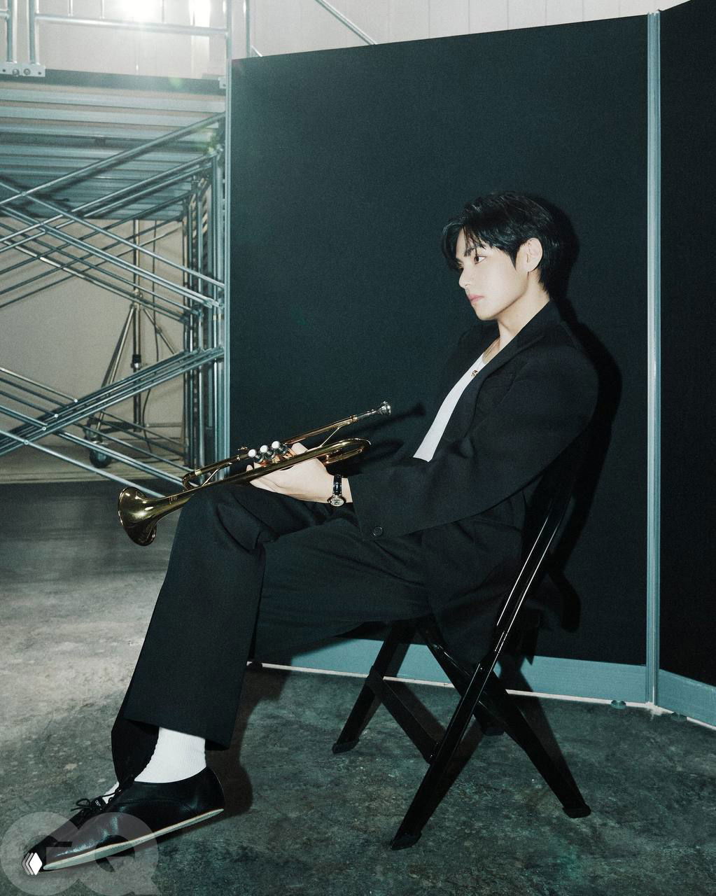 Фотография участника BTS, сидящего в кресле со струнным инструментом на фоне сценического реквизита — стильный backstage-портрет.