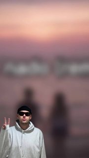 Размытое фото с человеком в кепке и очках в переднем плане, он показывает знак мира на фоне силуэта и мягкого заката — художественный фон снимка.