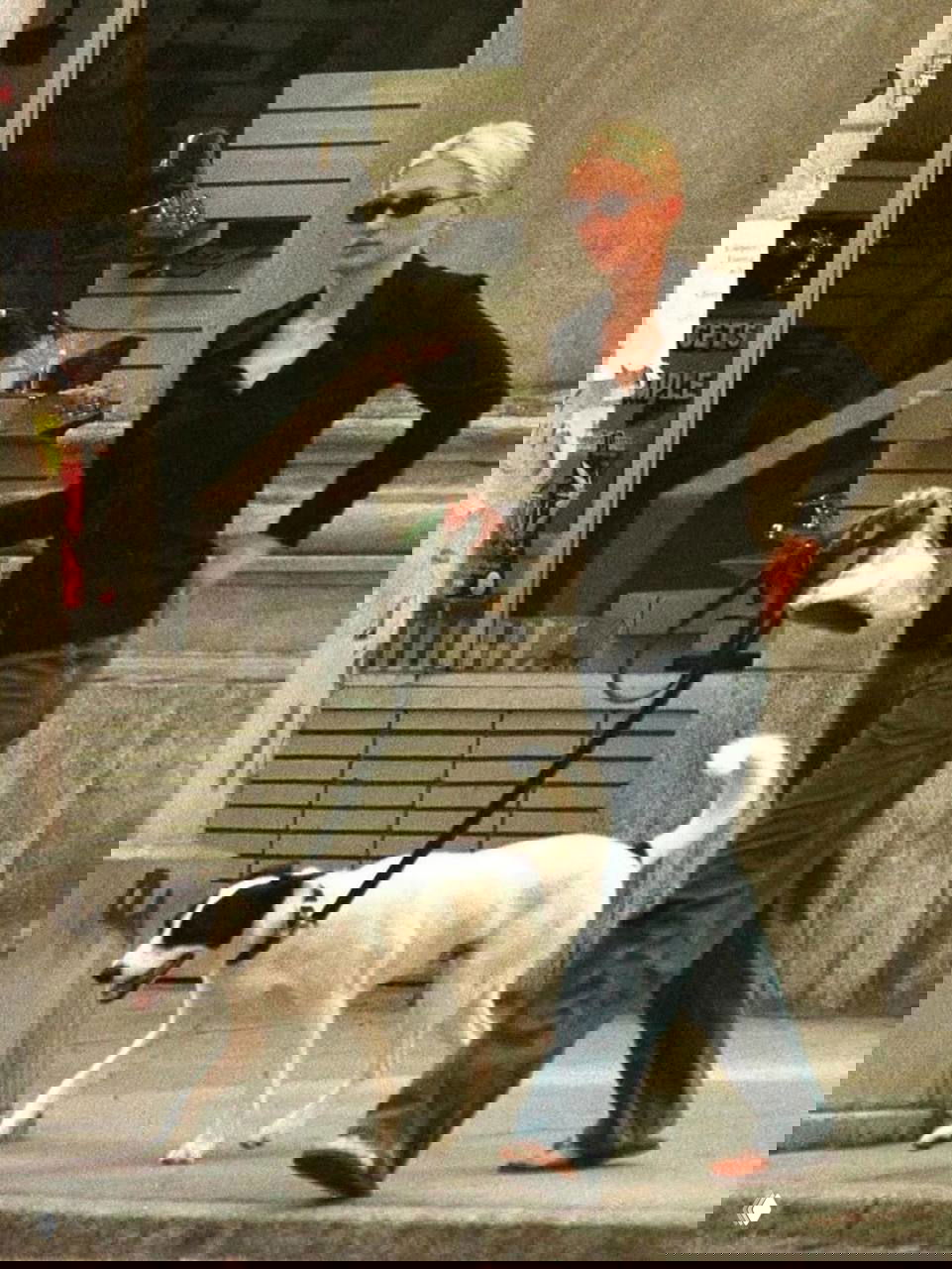 Женщина в черной рубашке и свободных джинсах идет с поводком, на прогулке с белой собакой в городской среде; кадр в стиле папарацци.