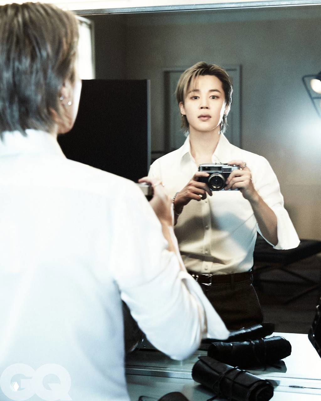 Участник BTS смотрит в зеркало, держит фотоаппарат — отражение и аккуратный образ в рубашке на съёмке для GQ.