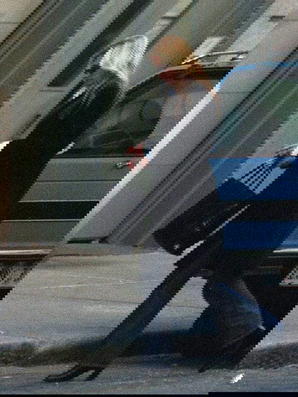 Женщина в длинном черном пальто и джинсах идет по тротуару, несет большую сумку; зернистая текстура кадра создает винтажную атмосферу.