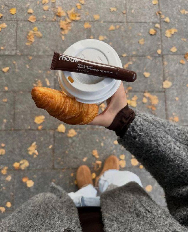 Снимок сверху: рука держит чашку кофе и круассан, на крышке лежит тюбик тинта для губ; осенние листья на тротуаре вокруг.