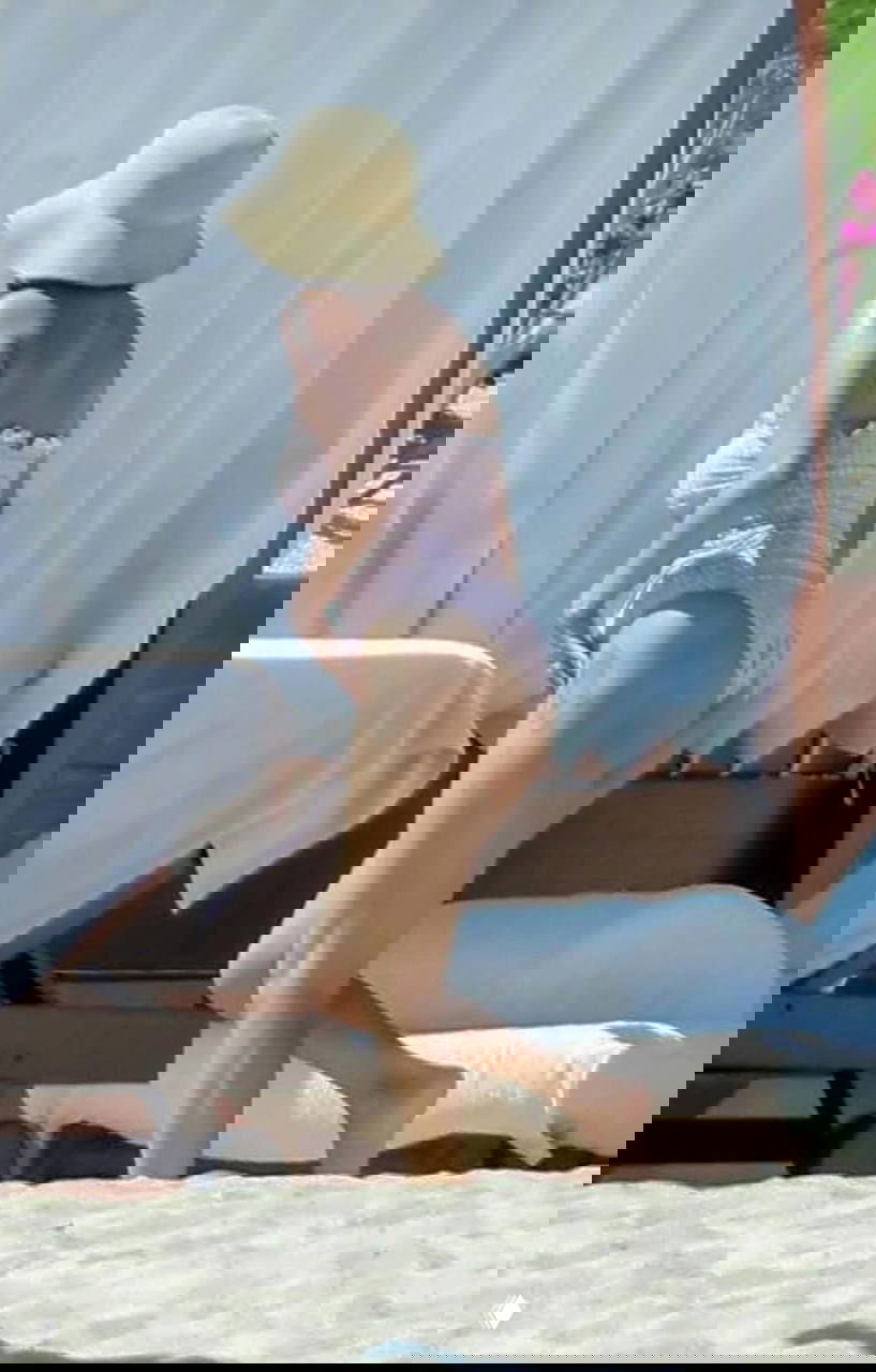 Селена Гомес в розовом купальнике и шляпе поднимается с пляжа у лежаков — кадр показывает позу и походку на песке во время отдыха.