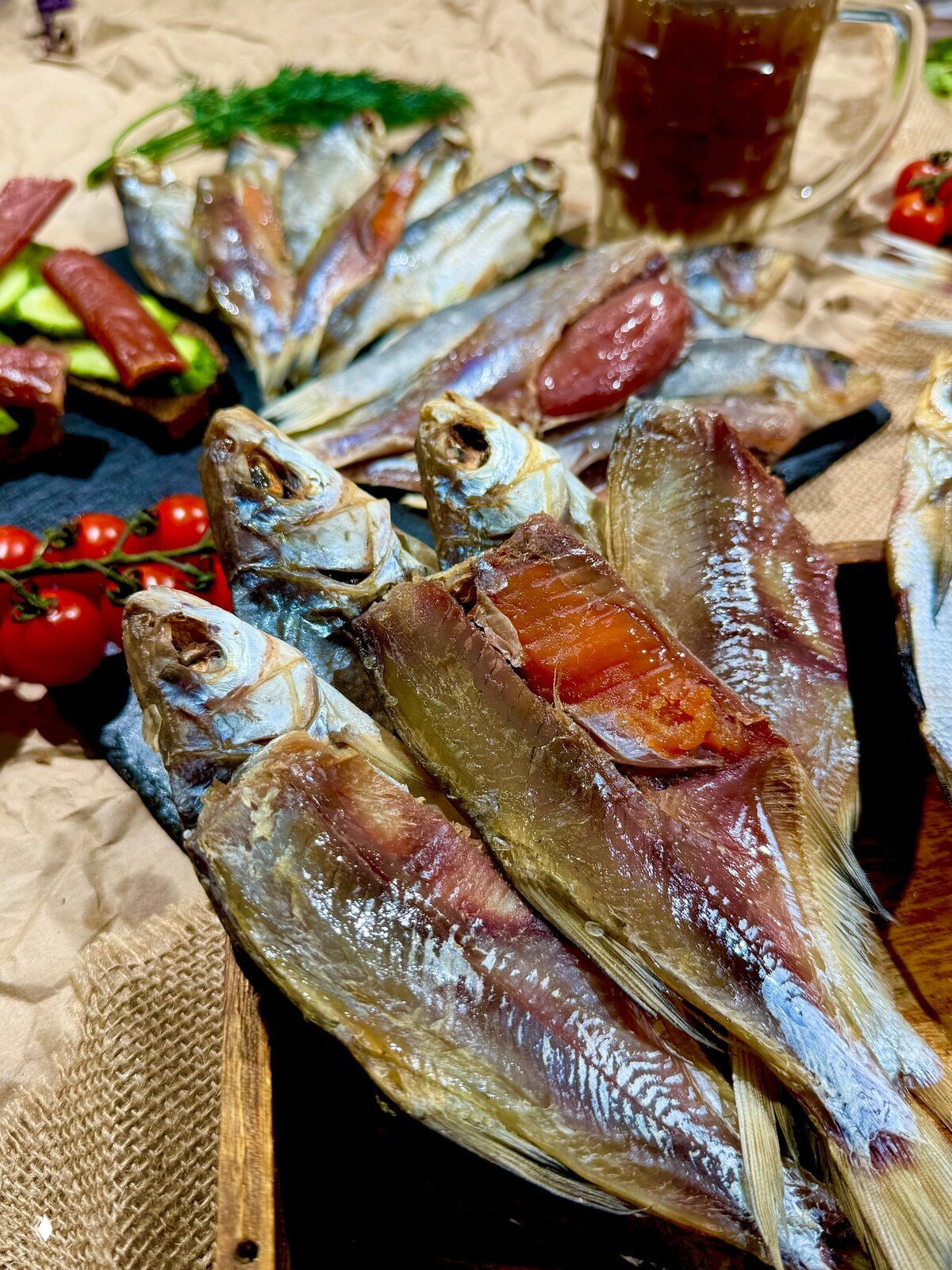 Крупный план вяленой рыбы на слэйте: видна блестящая кожа, мясистая текстура и фон с кружкой тёмного напитка и зеленью.