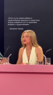 Крупный план женщины в белом топе за столом жюри на розовой сцене; микрофон и стакан воды перед ней, видно эмоциональное выражение.