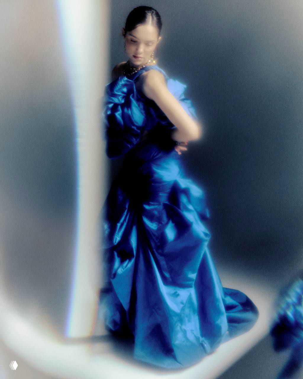 Размытый план в полный рост: женщина в элегантном синем платье со складками, скользящий свет и художественная зернистость кадра.