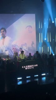 Вид сцены сбоку: диджей в белой рубашке, экран с проекцией сзади и скопление людей возле пульта в клубной обстановке.