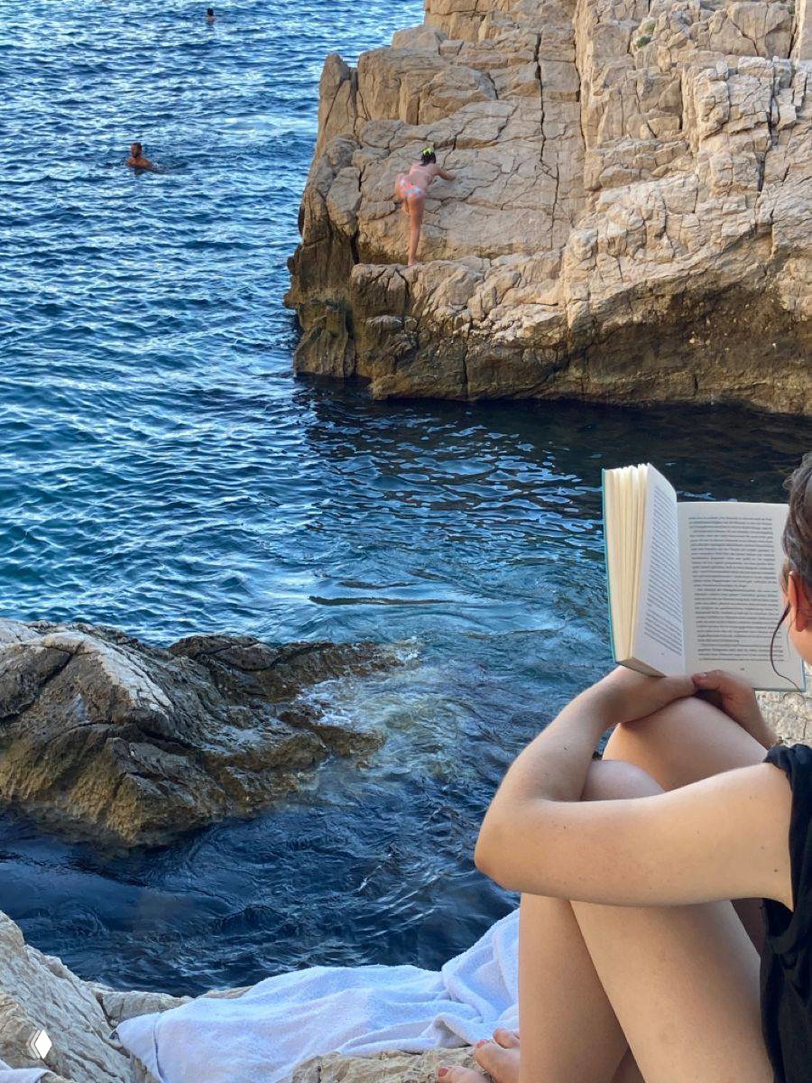 Человек сидит на скалах у моря и читает книгу; на заднем плане видны купающиеся и скалы, акцент на летнем чтении у воды