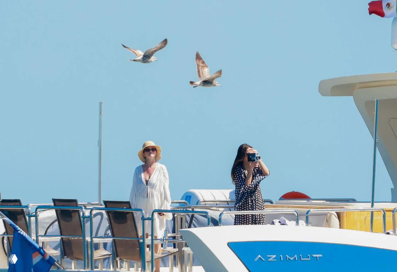 Широкий кадр яхты с Селеной Гомес и подругой, птицы в небе и открытое море на заднем плане — обширная сцена отдыха в Лос-Кабосе.