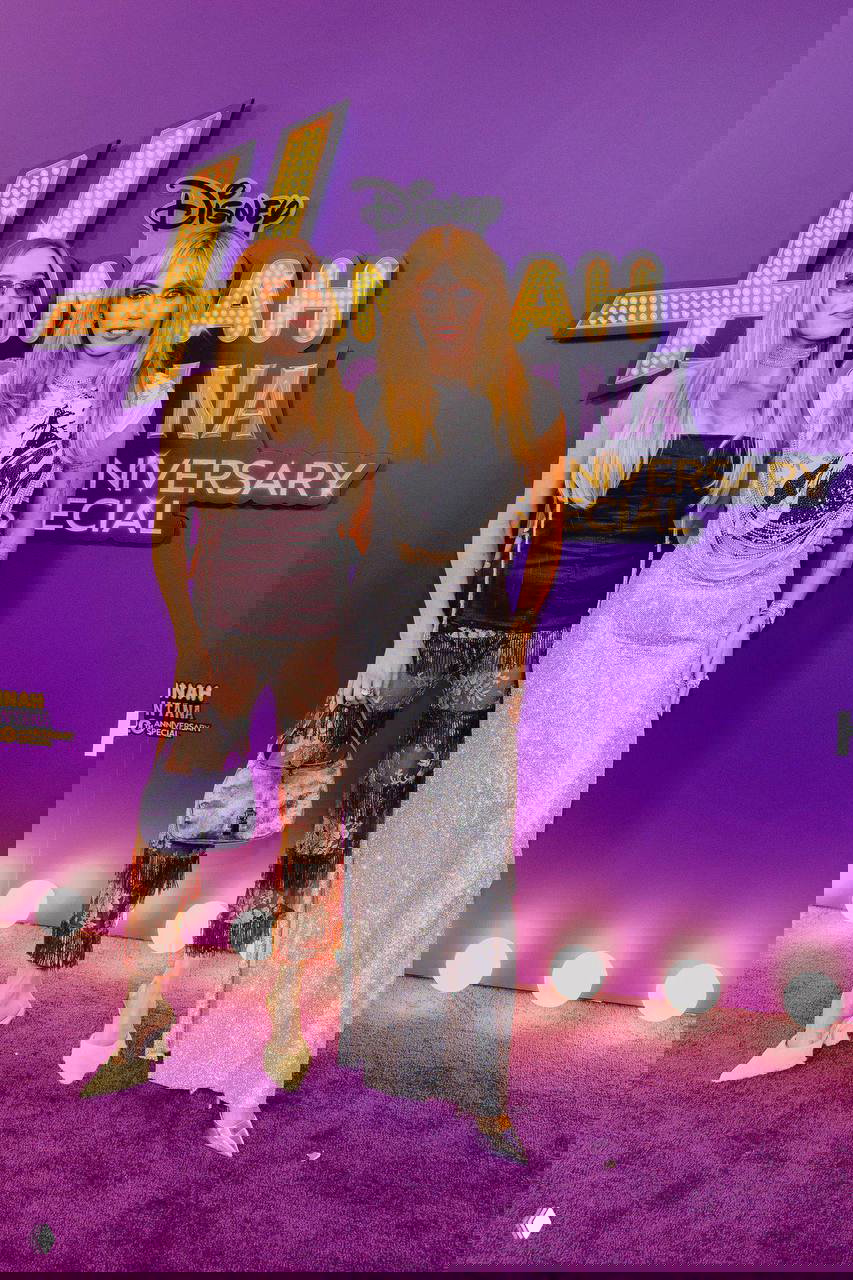 Майли Сайрус вместе с гостьей на фиолетовом фоне с логотипом «Hannah Montana», обе в вечерних образах — кадр с красной дорожки юбилея.