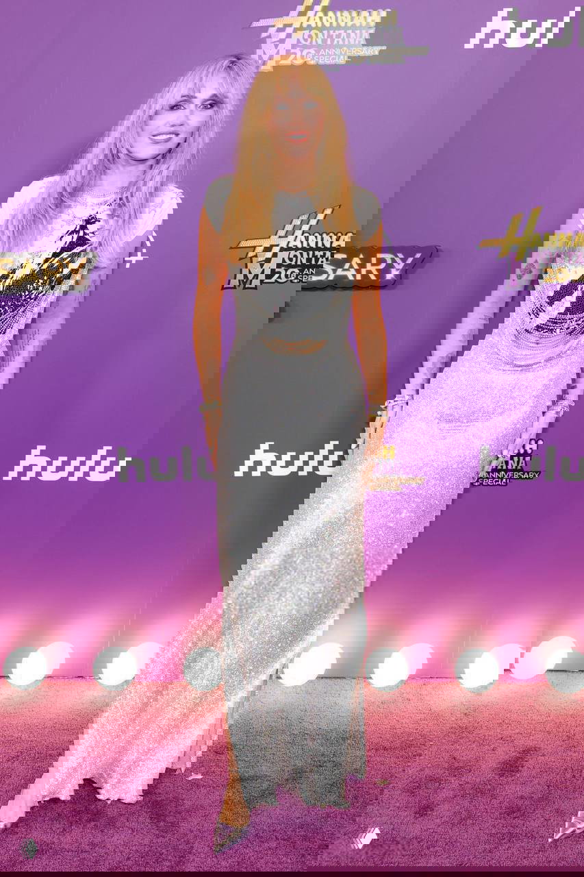 Полнотелый портрет Майли Сайрус в полный рост на фиолетовой ковровой дорожке юбилейного спецвыпуска «Ханна Монтана», серебристое платье и логотип позади.