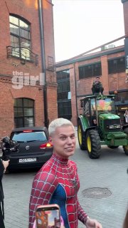 Человек в костюме Человека-паука на переднем плане, позади зелёный трактор и кирпичные фасады; логотип theGirl виден.