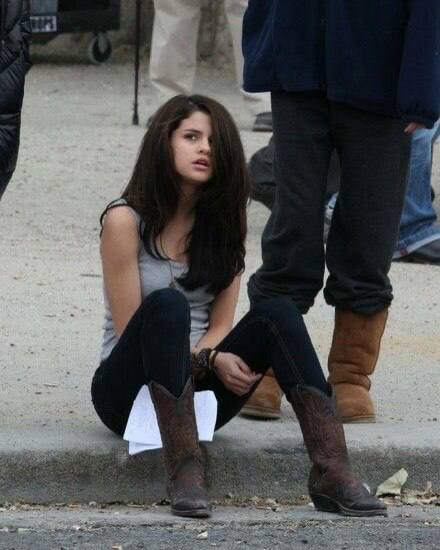 Селена Гомес сидит на тротуаре в 2010-х: длинные тёмные волосы, джинсы и ковбойские сапоги — уличный повседневный стиль папарацци.