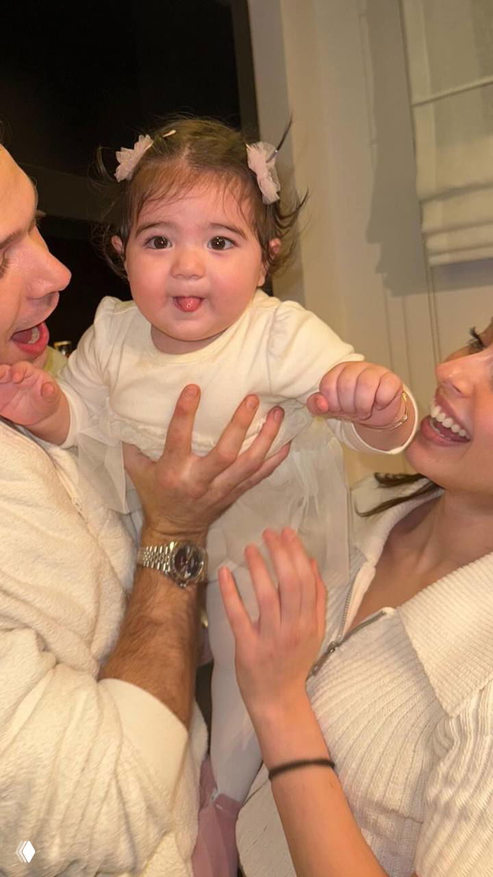 Джемре Байсел и Айтач Шашмаз поднимают ребёнка в кадре: белые вещи, улыбки и домашняя атмосфера, акцент на деталях образов.