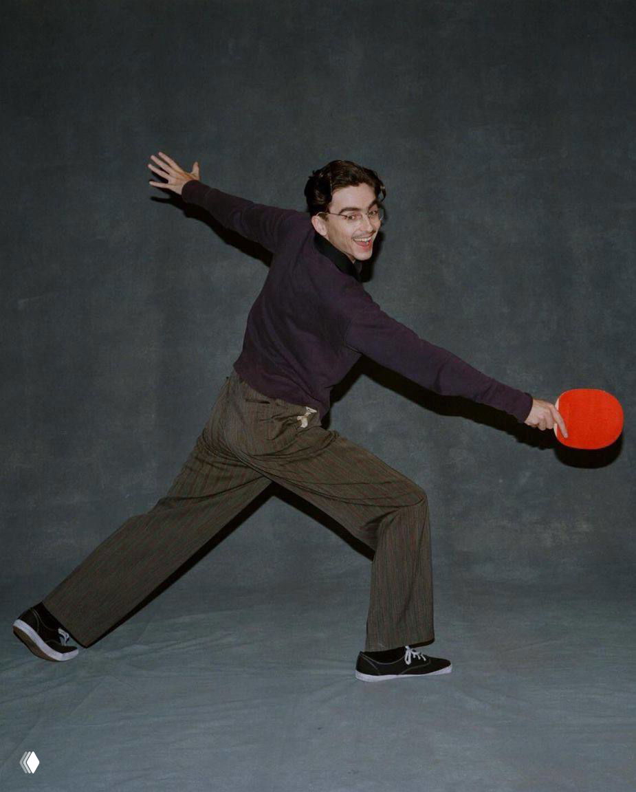 Фотография мужчины в динамичной позе с ракеткой для настольного тенниса на нейтральном фоне; игривый и энергичный промо‑кадр в студии.