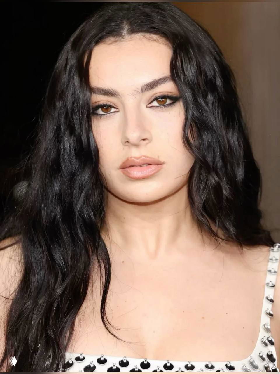 Портрет Чарли XCX: крупный план лица с тёмными волосами и выразительным макияжем, нейтральный фон, пресс-кадр.