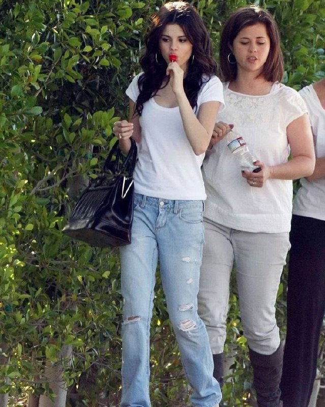 Селена Гомес в белой футболке и рваных джинсах с леденцом в руках, сопровождаемая помощницей — повседневный casual на прогулке.