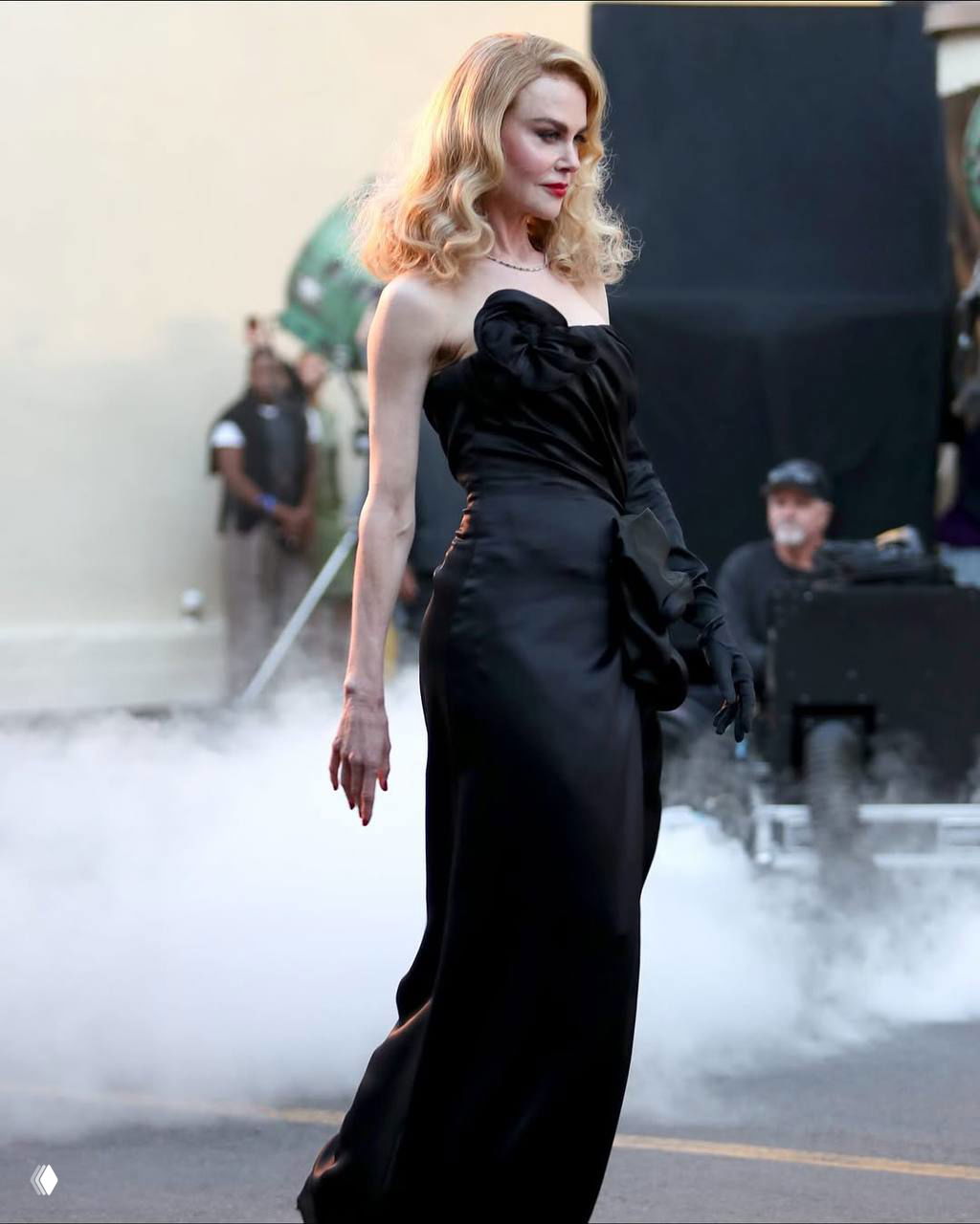 Знаменитость в элегантном длинном чёрном платье проходит мимо клубов дыма и съёмочной техники, создавая атмосферный кадр показа.