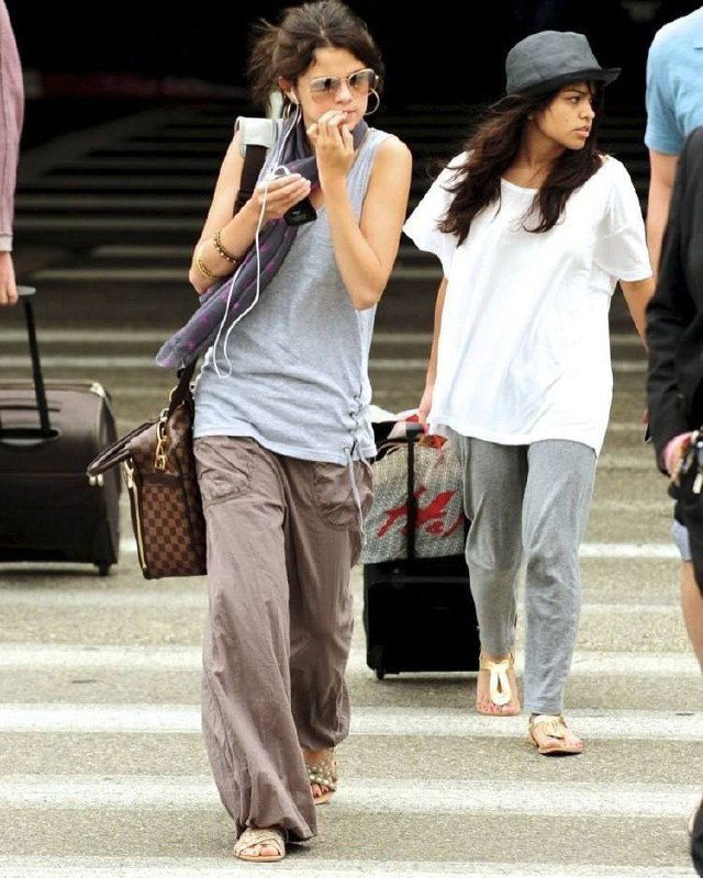 Молодая Селена Гомес в сером топе и широких брюках с сумкой и наушниками, в аэропорту — расслабленный travel‑лук 2010-х.