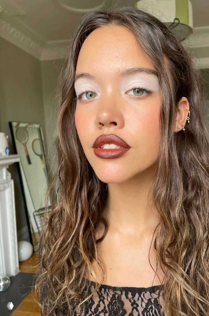 Крупный план молодой женщины с белыми тенями на веках и насыщенным цветом губ, пирсинг в носу и естественный интерьер на заднем плане.