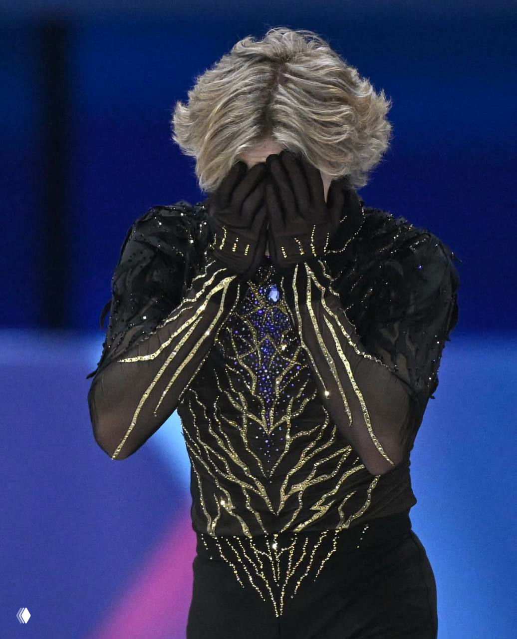 Фигурист в тёмном костюме закрывает лицо руками в эмоциональном кадре, видна отделка и декор костюма вблизи