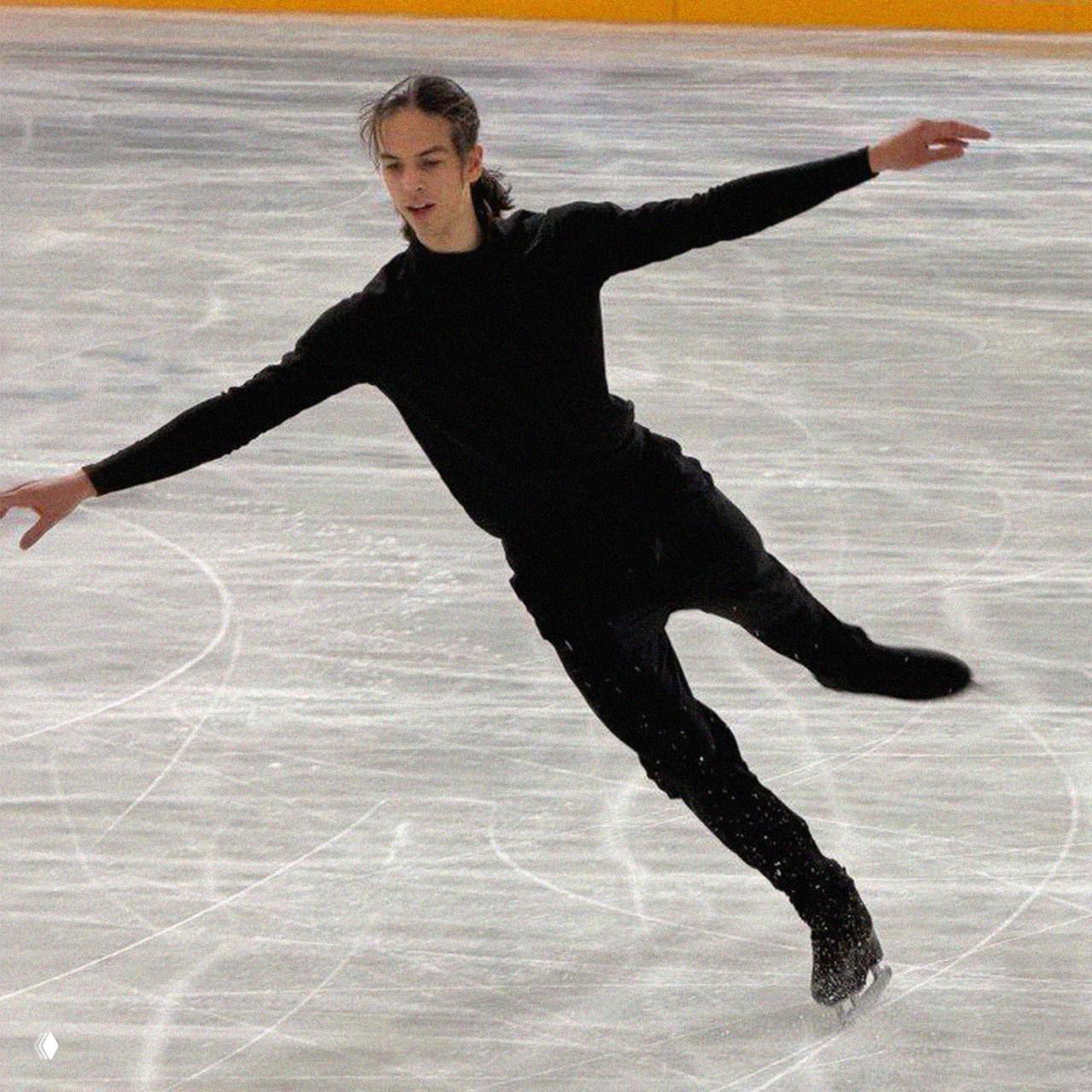 Фигурист в чёрном тренировочном костюме исполняет прокат на льду в прыжково-скоростном движении, видны следы от коньков на поверхности.