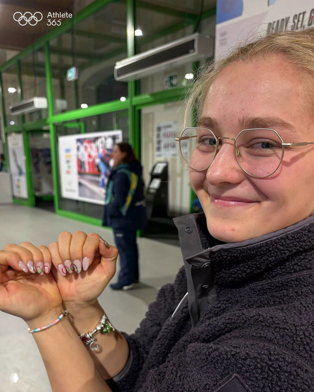 Спортсменка с очками показывает пастельный маникюр с тонкими рисунками и блестящими акцентами — фото в фойе олимпийского комплекса.