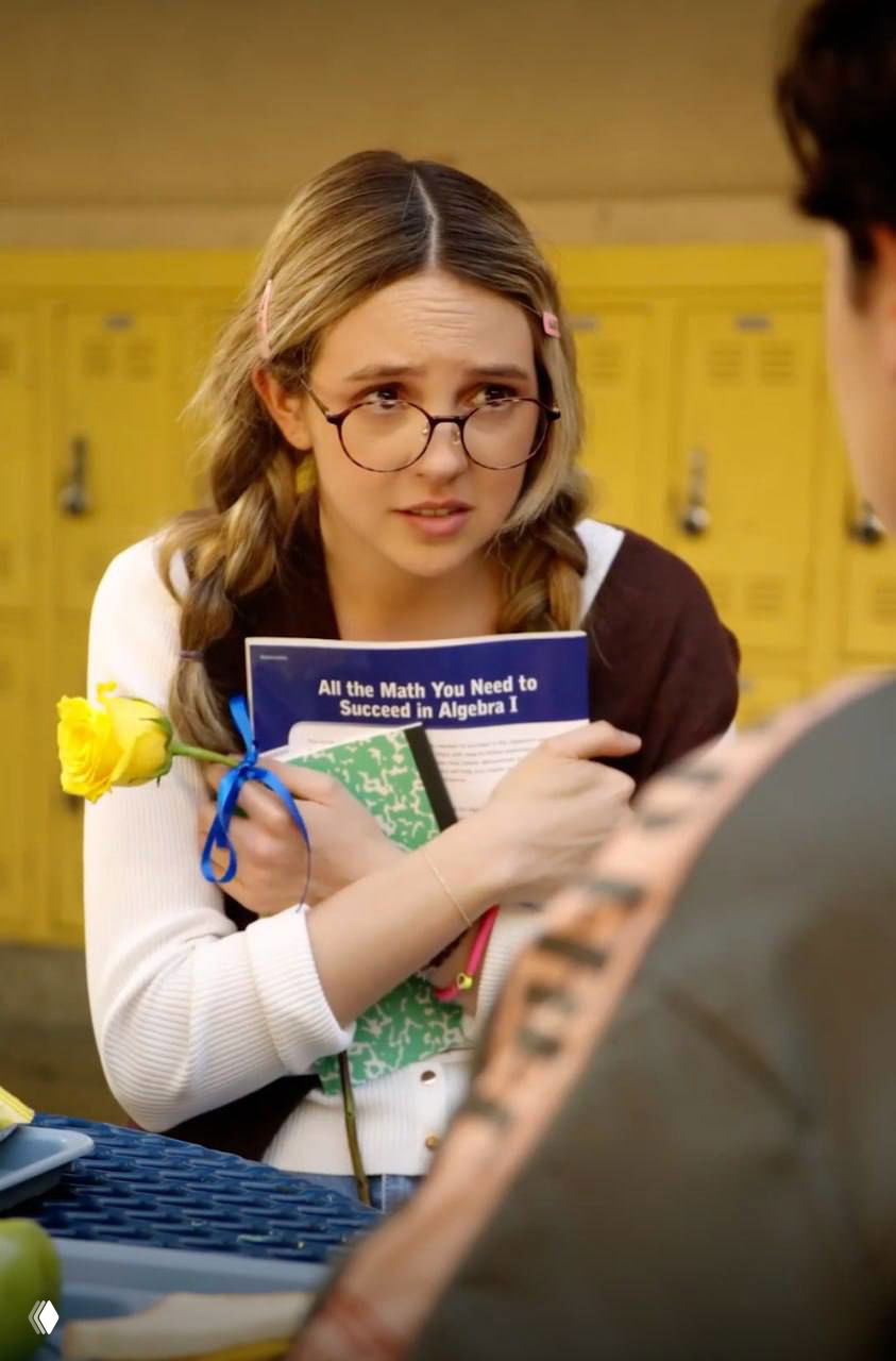 Девушка в очках держит учебник и жёлтый цветок в школьном коридоре; портрет юной героини из романтической мини‑драмы с подростковыми мотивами.