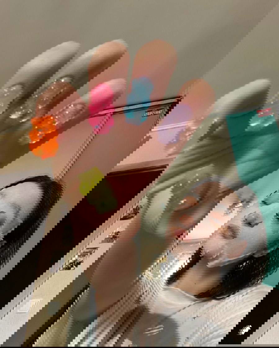 Рука с разноцветными желейными колечками на пальцах на фоне девушки в тканевой маске — сочетание ухода за кожей и мармеладных аксессуаров в молодежной эстетике.